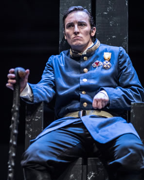 Richard sits on throne, frowning, left hand clinched with crooked arm, right hand on knotty cain, he's wearing a blue early 20th century dress uniform, long dress coat with gold-embroidered uptoruned collar, gold buttons and belt, and two medals on his left breast